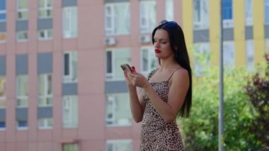 Leopar desenli elbiseli bir kadın apartmanın yakınındaki akıllı telefonunu kontrol ediyor..