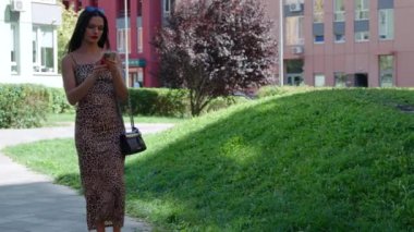 Bir kadın şehir parkında şık bir leopar desenli elbiseyle dolaşır ve akıllı telefonuna odaklanır..
