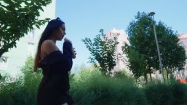 Sıcak bir öğleden sonra yemyeşil bir şehir ortamında yemyeşil bir ortamda duran genç bir kadın içkisini yudumluyor..