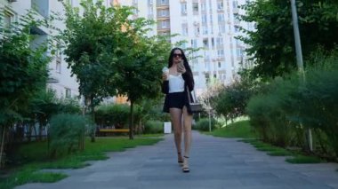 Zarif bir kadın elinde bir fincan kahveyle yaz modasına uygun bir şekilde lüks bir parkta geziniyor..