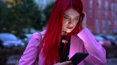 Kızıl saçlı genç bir kadın dışarıda dikilirken akıllı telefonuna odaklanmış..