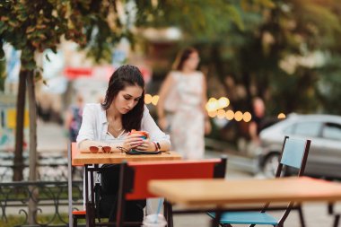Kafenin dışındaki bir masada oturan bir kadın, akıllı telefonuna odaklanmış, çevresinin tadını çıkarıyor..