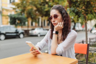 Kafe masasında oturan genç bir kadın, düşünceye dalmışken akıllı telefonunu kontrol ediyor..