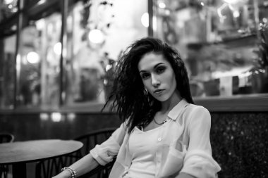 Kıvırcık saçlı genç bir kadın şık bir kafede oturmuş, sıcak ışıklarla süslenmiş..