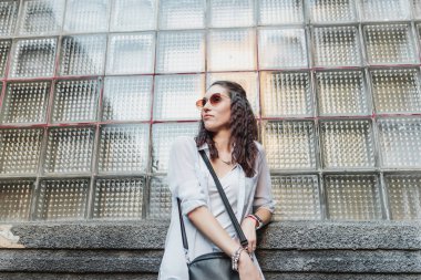 Genç bir kadın, modaya uygun güneş gözlüğü takarken gelişigüzel dokulu bir duvara yaslanıyor..