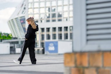 Genç bir kadın, berrak gökyüzünün altında çağdaş bir şehir ortamında gelişigüzel dolaşıyor..