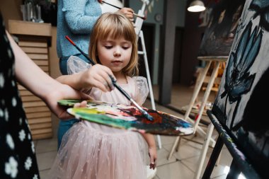 Elbise giymiş küçük bir kız elinde paletle tuvale resim yapıyor..