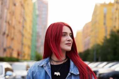 Uzun kızıl saçlı genç bir kadın hayat dolu bir şehirde gülümsüyor..