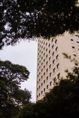 Bulutlu bir gökyüzünün altında yemyeşil ağaçların çerçevelediği yüksek bir binayı gösteren bir manzara..