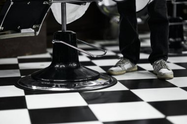 Bir berber kareli zeminde şık siyah bir berber sandalyesinin yanında duruyor..