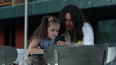 Bir anne, dışarıda otururken kızına akıllı telefondan bir şey gösterir..