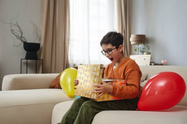 Genç bir çocuk sevinçle hediyesini açar ve balonlar yakınlarda yüzer..