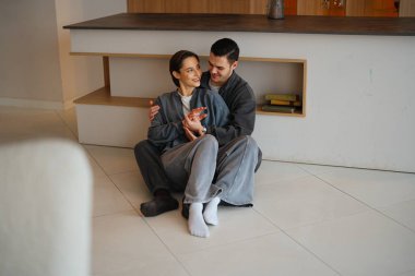 Bir çift yerde oturuyor, gülümsüyor ve evde sıcak bir anı paylaşıyor..