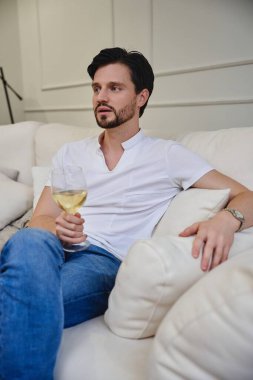 Genç bir adam rahat bir şekilde kanepede oturup bir bardak beyaz şarabın tadını çıkarıyor..