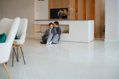 Çift yerde oturuyor, modern bir yaşam alanında birbirlerine sarılıyorlar..