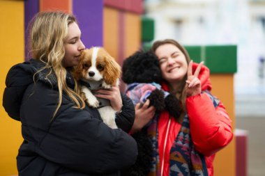 İki genç kadın canlı ve renkli bir ortamda küçük köpekleriyle neşe içinde poz veriyorlar. Bir kadın köpeğini öperken diğeri gülümser ve barış işareti yapar. Eğlenceli bir ortam yaratır..