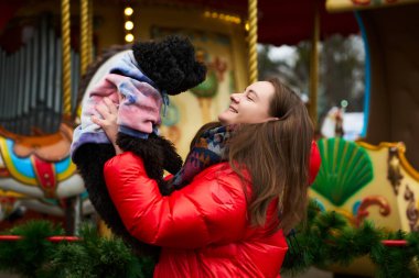 Kırmızı ceketli bir kadın siyah köpeğini renkli bir kazakla tutarken parlak bir şekilde gülümsüyor. Bayram süslemeleriyle çevrili canlı bir atlıkarınca yakınlarında kış şenliklerinin tadını çıkarıyorlar..