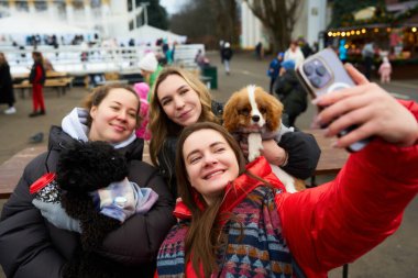 Canlı bir parkta toplanan üç arkadaş sevimli köpekleriyle selfie çekerken gülümsüyorlar. Neşeli bir kış gününün altını çizerek onları çevreleyen neşeli atmosfer.