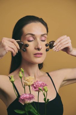 Bir kadın elinde çiçeklerle bir stüdyoda yüzüne güzellik aletleri uyguluyor..