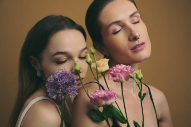 Gözleri kapalı iki kadın ellerinde çiçeklerle güzelliklerini sergiliyorlar..