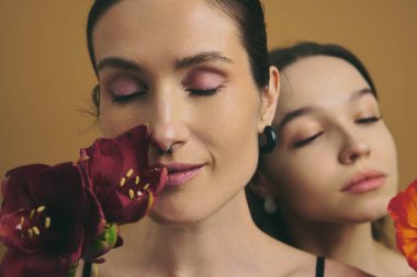 İki kadın, sükuneti kucaklarken ellerinde çiçeklerle birbirlerine yakın dururlar..