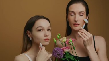 İki kadın stüdyo ortamında çiçek kullanarak cilt bakımı teknikleri gösteriyor..