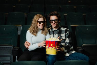 Patlamış mısır paylaşırken gülümseyen, sinema koltuklarında 3D gözlük takan bir çift..