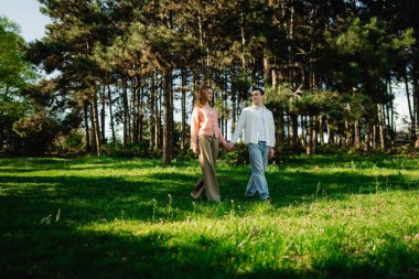 Ağaçların arasında yeşil çimenlerde yürürken iki insan el ele tutuşur..