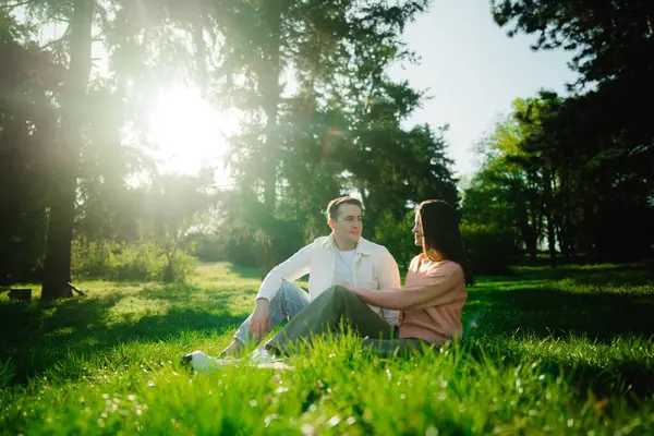 İki insan yemyeşil ağaçlar ve güneş ışığıyla çevrili çimenli bir yüzeyde rahatlar..