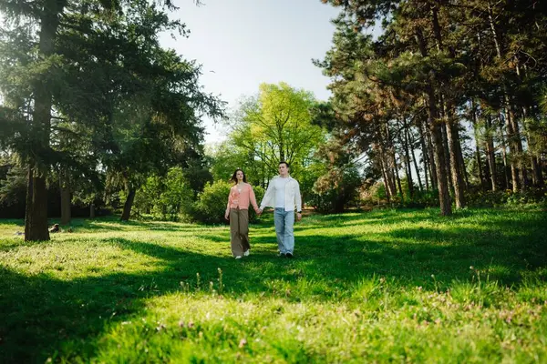 İki birey, açık mavi gökyüzünün altında canlı çimlerin üzerinde el ele yürüyor..