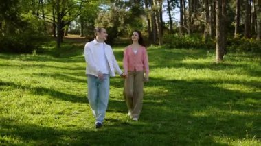 Genç bir çift, güneşin altındaki yeşil bir parkta el ele dolaşır..