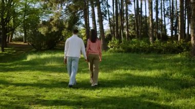 Bir çift el ele yürüyor, yemyeşil ve uzun ağaçlarla çevrili..