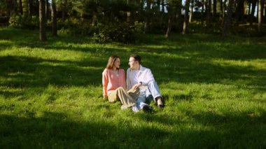 İki insan, ağaçların altındaki yeşil çimlerde otururken huzurlu bir anı paylaşıyor..