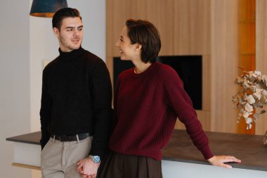 Genç çift şık bir mutfakta dururken birbirlerine gülümsüyor..