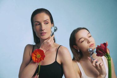 İki kadın yan yana durur, yüz gereçleri kullanır ve ellerinde renkli çiçekler tutarlar..