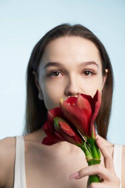 Genç bir kadın sakince yüzünün yanında kırmızı çiçekleri tutuyor..