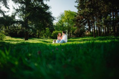 Parlak güneş ışığı altında ağaçlarla çevrili yeşil çimlerde iki insan rahatlar..