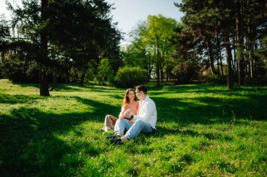 Yumuşak çimenlerde oturan gülümseyen bir çift, aydınlık bir günde ağaçlarla çevrili..