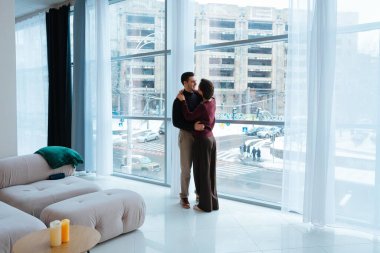 Caddeye bakan büyük pencerelerin yanında duran iki kişi sarılıyor..