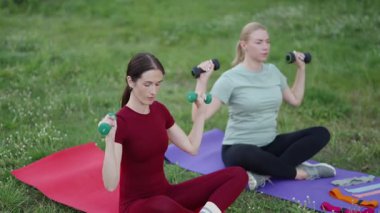 İki kadın dışarıda spor yapıyor, yeşil park ortamında ağırlık kaldırıyor..