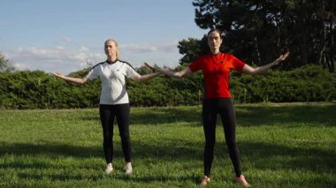 İki kadın açık bir gökyüzünün altındaki yeşil bir parkta senkronize ısınma egzersizleri yapıyor..