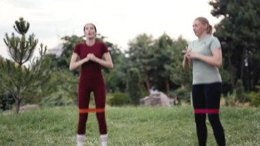İki kadın parkta spor yapıyor. Formlarını arttırmak için direnç bantları kullanıyorlar..