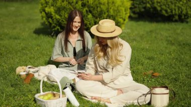 İki arkadaş parkta bir battaniyenin üzerinde gevşeyip güneşin altında çerezleri ve kahkahaları paylaşıyorlar..