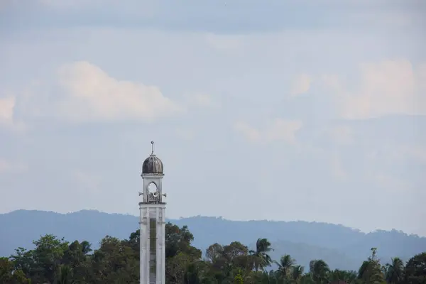 Bir cami minaresi, yemyeşil yemyeşil bir zemin ve dağınık bulutlu sakin bir gökyüzünün altında uzak dağlara karşı dimdik ayakta duruyor. Seyahat, mimari ve doğa için mükemmel..