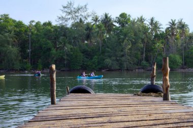 Kırsal bir ahşap iskele sakin bir tropikal nehre doğru uzanır. Kayıkçılar palmiye ağaçlarının yemyeşil zeminine ve sık ormanlara karşı kürek çeker.. 