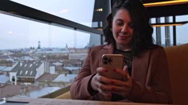 Kahvede oturan gülümseyen kadın telefondaki haberleri kontrol ediyor.