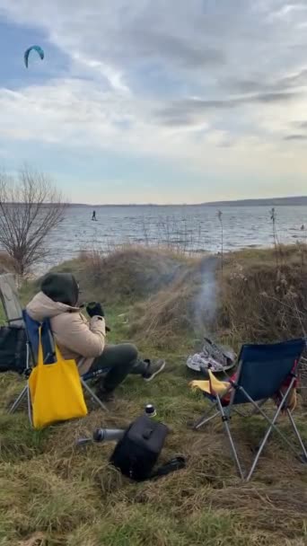 Mężczyzna Pijący Herbatę Pobliżu Bonfire Windy Day Kitesurfers Nad Jeziorem — Wideo stockowe