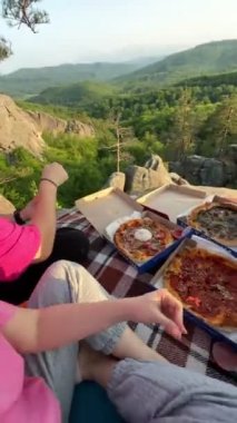 Mutlu arkadaşlar uçurumda pizza yerken gün batımı yürüyüşü konseptine bakarlar.