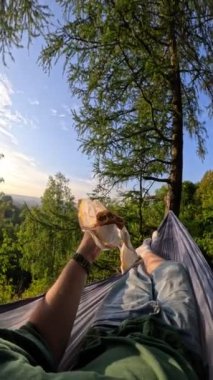 Pov adam hamakta uzanıp hamburger yiyor ve günbatımının tadını çıkarıyor.