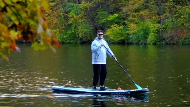 秋の湖の真ん中にあるスーパーボードの男の景色 背景にカラフルな森 — ストック動画
